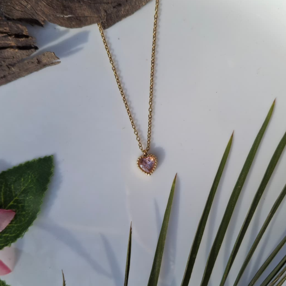 Necklace with a pink heart-shaped pendant on a white surface with green leaves and brown wood.