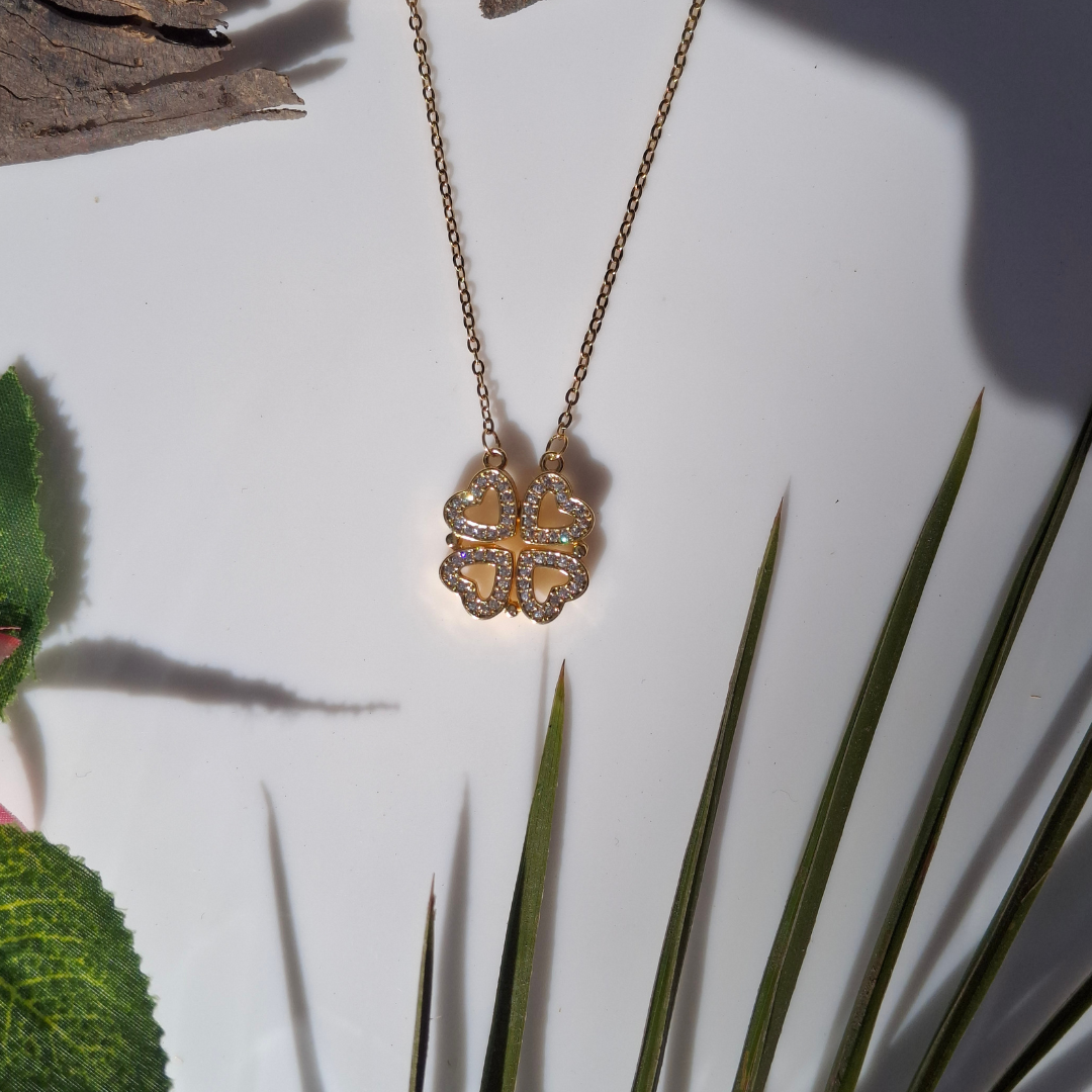 Gold necklace with a clover-shaped pendant on a white background with green leaves.